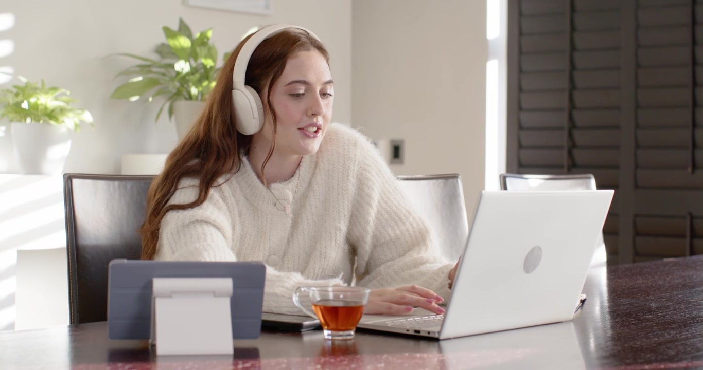 Adult woman wearing white headphones and cream sweater typing and speaking to laptop, tea visible