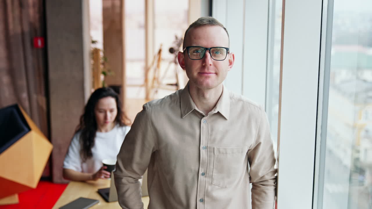 Mid-aged man wearing glasses stands at window. Man in shirt smiles to camera. Woman at backdrop drinking coffee working at laptop.