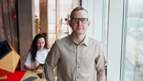 mid-aged man wearing glasses stands at window. man in shirt smiles to camera. woman at backdrop drinking coffee working at laptop.