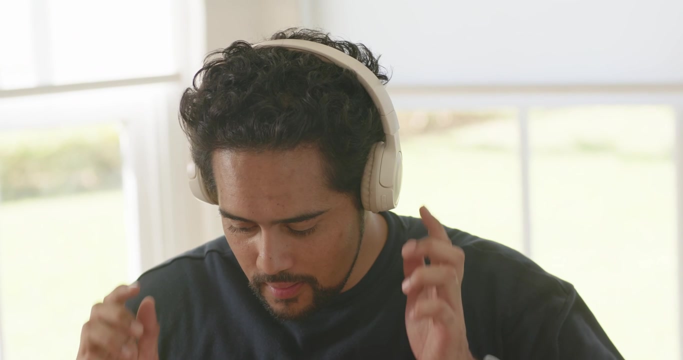 Uomo africano di mezza età che afferra cuffie over-ear bianche, regolandole vicino a una finestra per ascoltare.