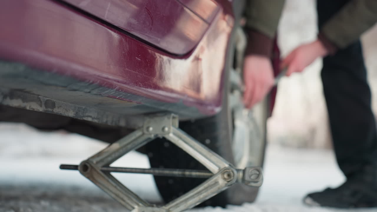 Close up of car jack positioned under vehicle on snowy ground, person slightly loosening bolt on rim, clear focus on tool movement and tire, cold outdoor setting with focus on mechanical work