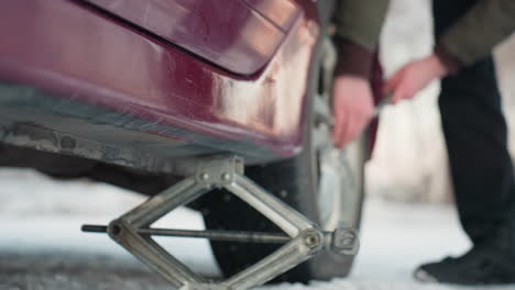 close up of car jack positioned under vehicle on snowy ground, person slightly loosening bolt on rim, clear focus on tool movement and tire, cold outdoor setting with focus on mechanical work
