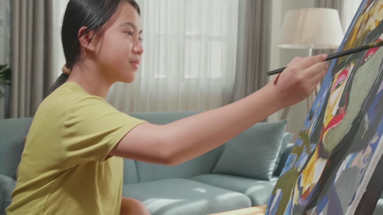 Side View Of An Asian Artist Girl In Wheelchair Holding Paintbrush Mixed Colour And Painting A Girl On The Canvas