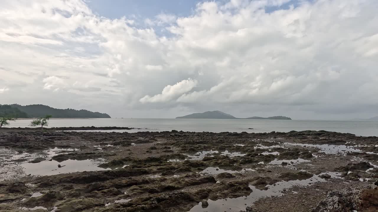 A slow camera pan reveals a rocky coastal shoreline, sandy beach, and lush forest under dramatic cloudy skies at Ko Phayam, Thailand