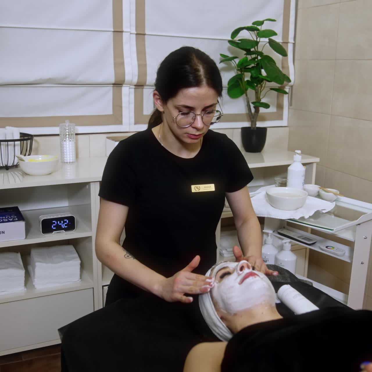 Female cosmetologist applies mask on the client's face. Beauty specialist carefully performs massage to a woman lying on the couch