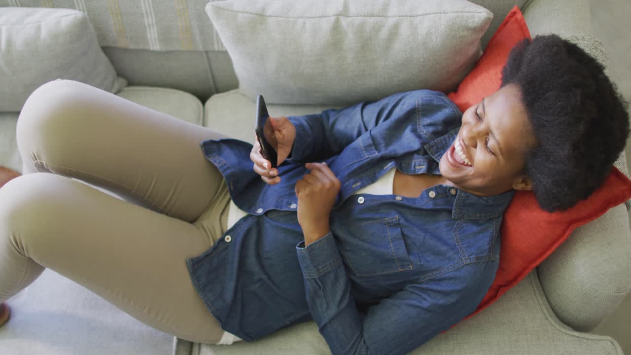 Happy african american woman laying on sofa, using smartphone