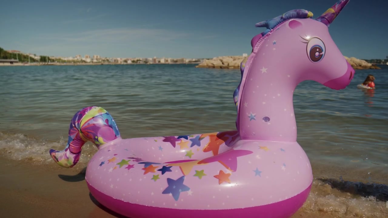 Phao bơi bơm hơi hình kỳ lân màu hồng nằm trên bờ cát của bãi biển Plage de la Gravette ở Antibes, Pháp, với làn nước Địa Trung Hải tĩnh lặng và đê chắn sóng đá ở phía sau