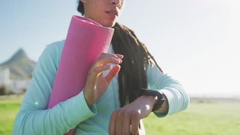 african american woman in sportswear holding mat using smartwatch in park