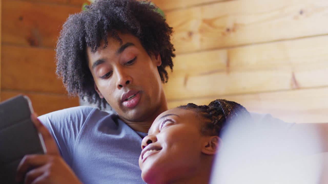 Coppia afroamericana felice che usa il tablet, seduta sul divano in una baita di legno, al rallentatore