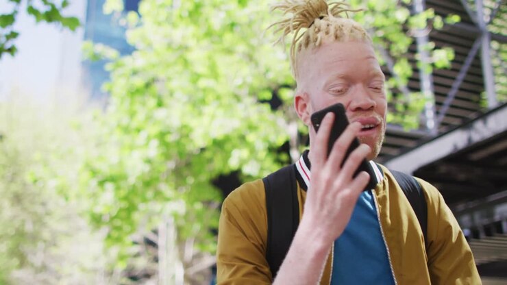 Glad albino afroamerikansk mand med dreadlocks i parken, der taler i smartphone