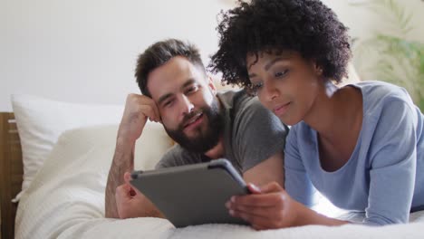 video of happy diverse couple relaxing at home, lying on bed using tablet