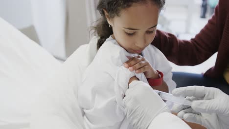 doctor vaccinating biracial sick girl patient with mother in hospital in slow motion