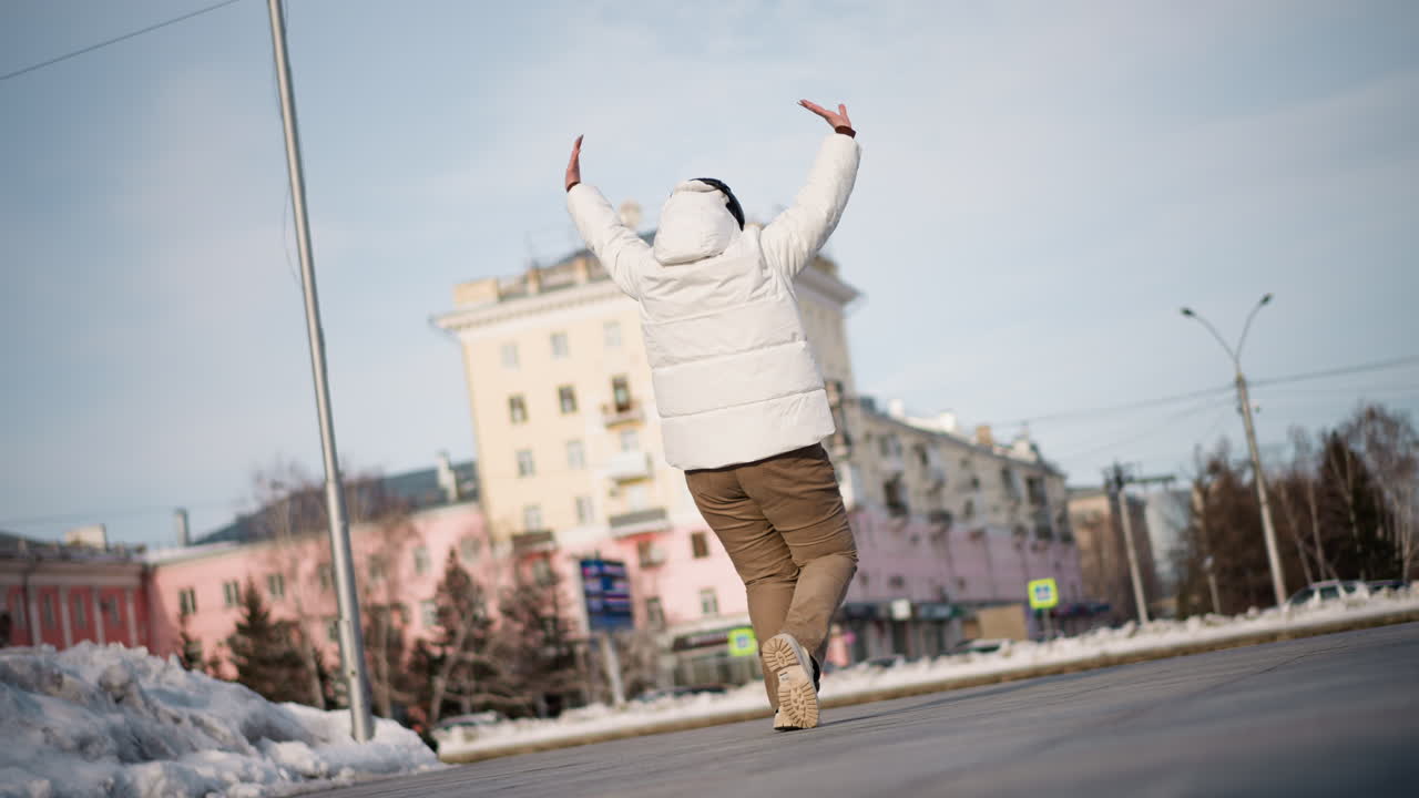 Female figure swinging arms and stepping gracefully across snowy urban plaza, wearing white puffer jacket, beanie and headphones, expressing emotion through fluid dance under pale sky