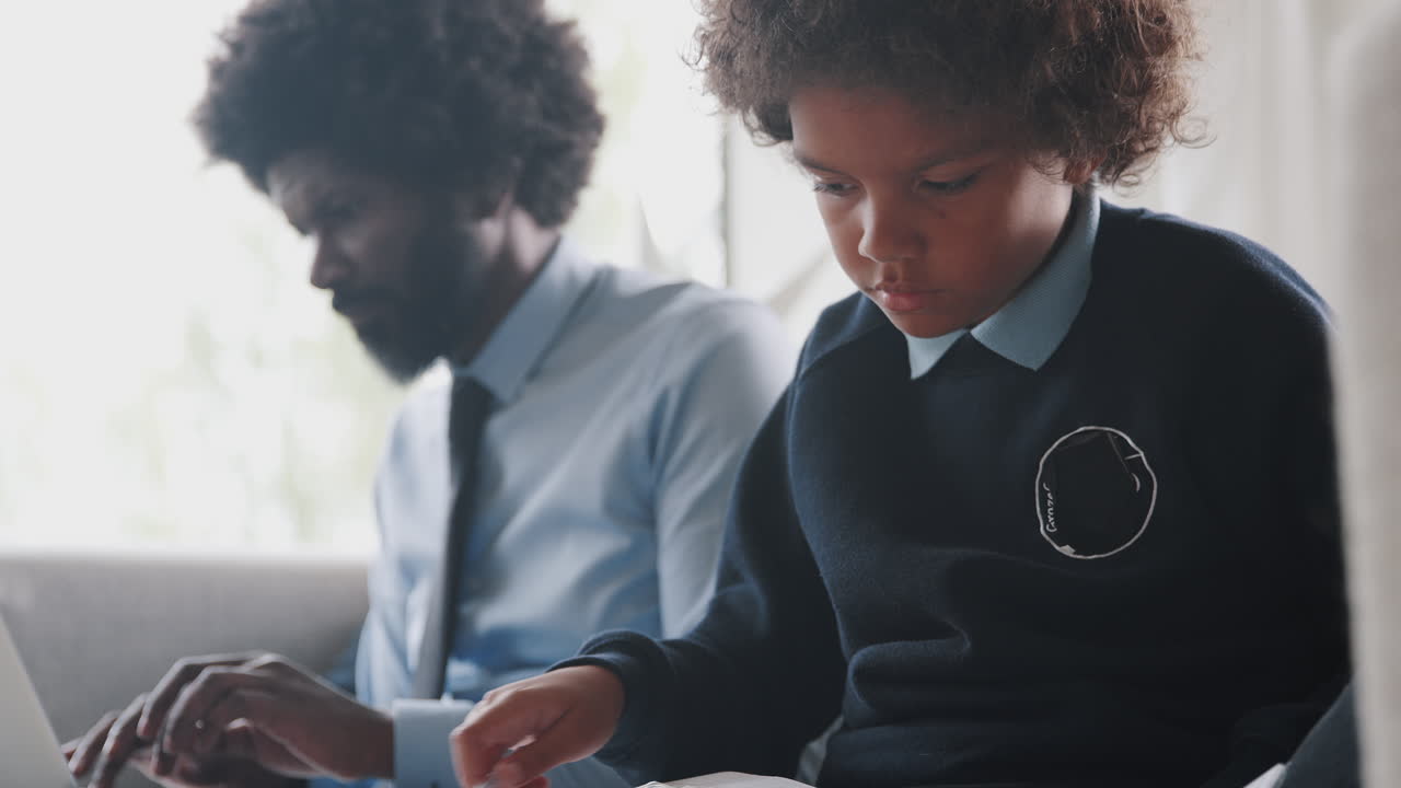 Close up of pre-teen schoolboy sitting on the sofa at home doing homework and his father sitting beside him, focus on foreground