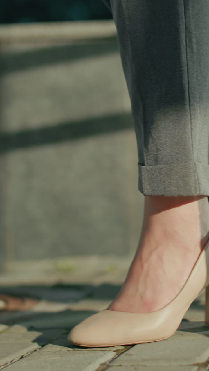 Leg view of young woman walking on interlocked pavement wearing beige heels and grey trousers, sunlight casting shadows along sidewalk with stone wall and urban railings in background