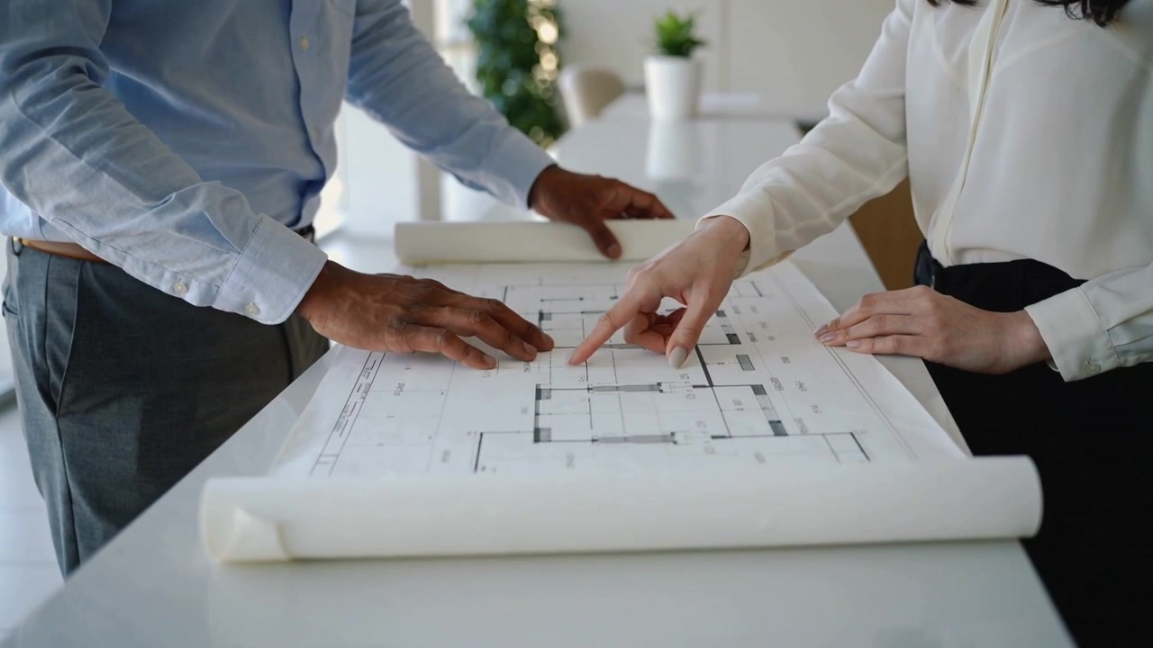 Unfurling plan, blue-shirt man and white-blouse woman pointing, reviewing layout at office table