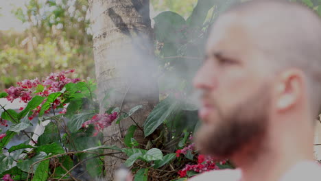 green vegetation and red flowers in the background, in front of a defocused bearded bald man smoking vape