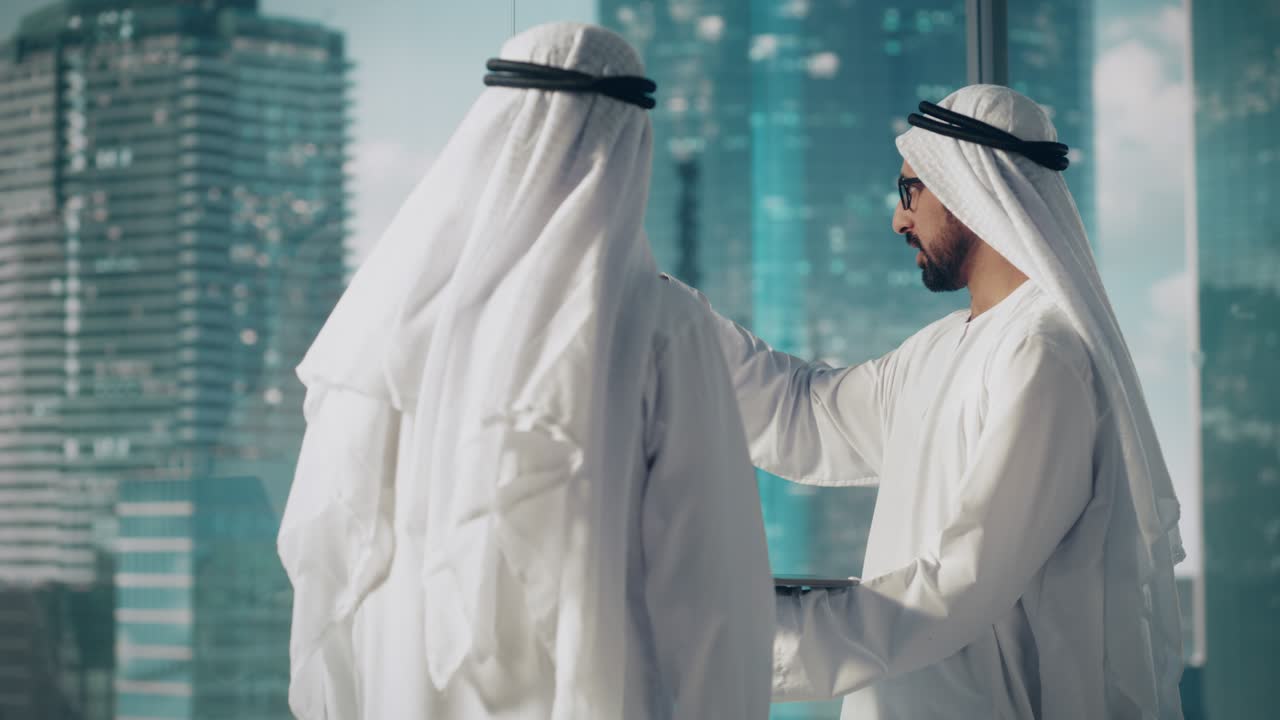 due uomini d'affari musulmani di successo in abiti tradizionali bianchi in piedi in ufficio che si incontrano, negoziano e parlano di opportunità finanziarie, usando un portatile.