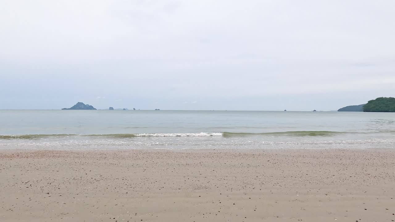 Gentle waves roll onto the sandy shore of Nopparat Thara Beach, Krabi, under overcast skies, creating a tranquil seaside scene