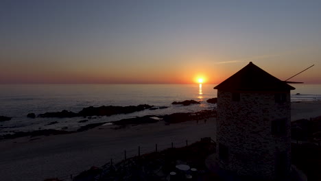 Toma-Aérea-Panorámica-De-4k-De-Un-Molino-De-Viento-Como-Punto-De-Interés-Cerca-De-La-Costa-Del-Mar-Que-Revela-Una-Hermosa-Puesta-De-Sol