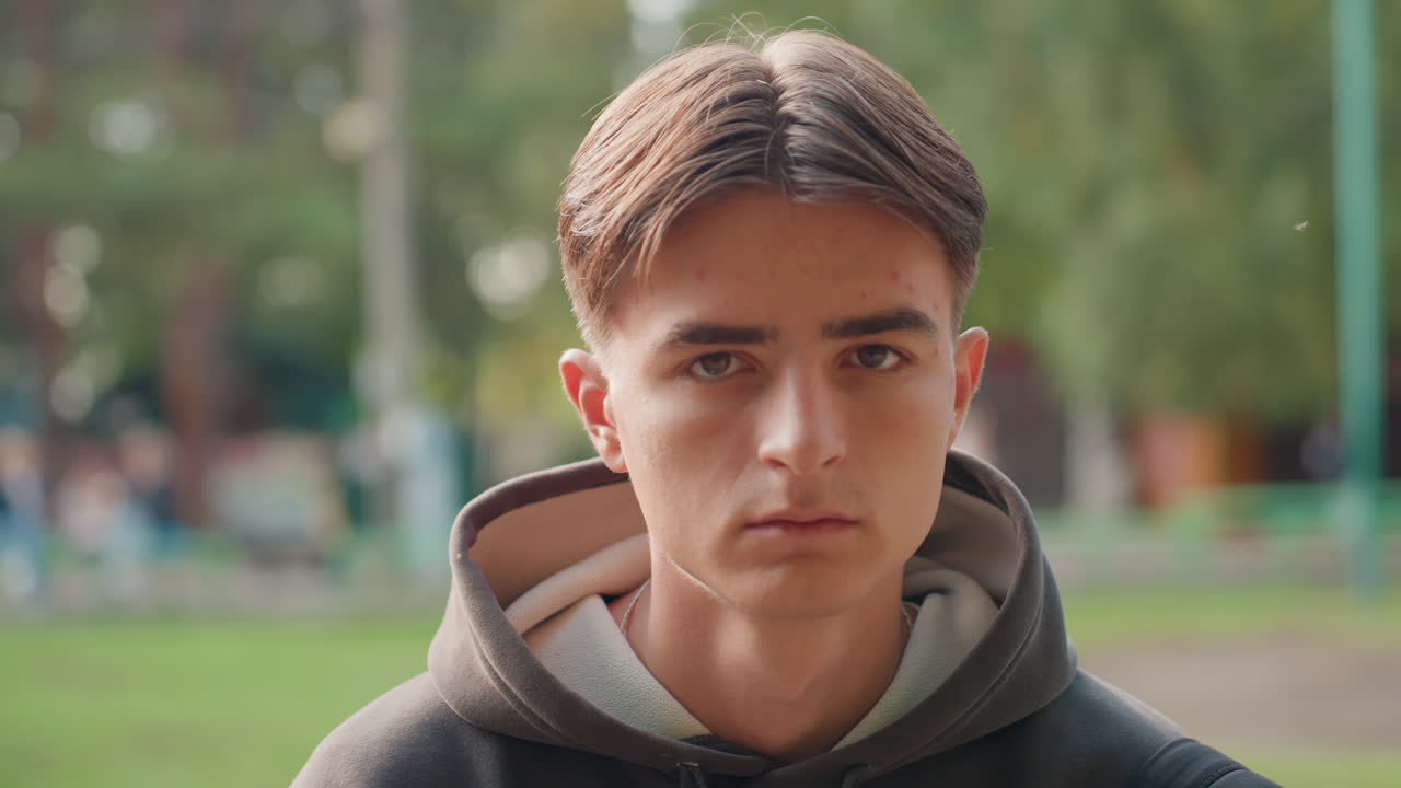 Giovane uomo serio con felpa, primo piano in un parco che mostra uno sguardo determinato e un leggero effetto bokeh degli alberi, luce naturale sul viso, studente che contempla i prossimi passi con la borsa di lato, atleta che si rilassa dopo l'allenamento
