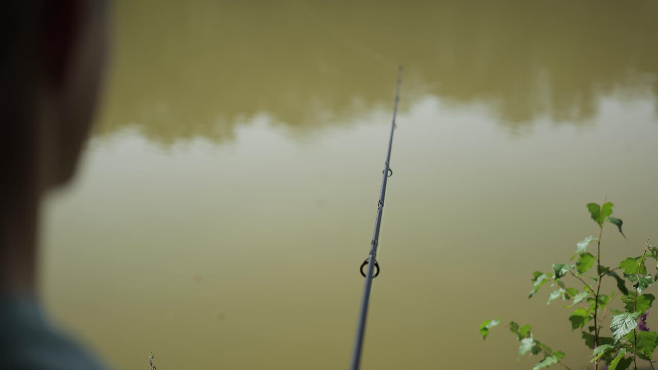Male angler stands on the shore of a very cloudy murky lake and holds fishing rod into the water