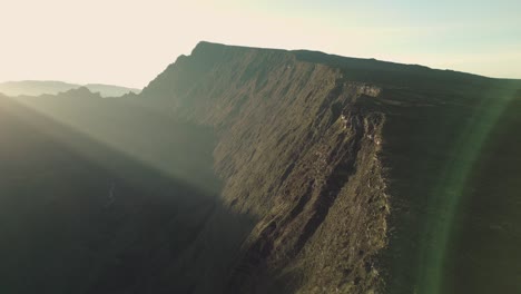 Drone-flying-slowly-along-a-huge-crater-wall-at-sunrise-with-sun-flares-over-Mafate-in-La-Reunion