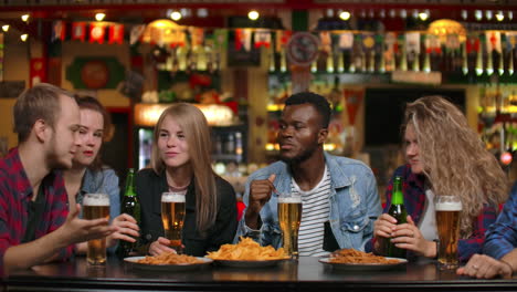 amigos multiétnicos de grappa sentados en un bar bebiendo cerveza y divirtiéndose charlando mientras comen papas fritas.