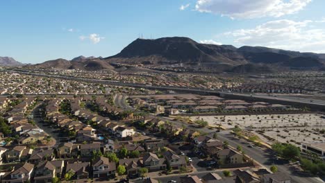 Drone-Ascendiendo-Sobre-Los-Suburbios-En-Henderson-Nevada-Con-Carretera-Y-Montañas-En-El-Cielo-Azul-De-Fondo-60fps