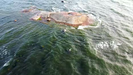 Una-Suave-Toma-Aérea-De-Un-Lago-Que-Fluye-Y-Olas-Golpeando-Una-Pequeña-Isla-Rocosa-En-La-Superficie