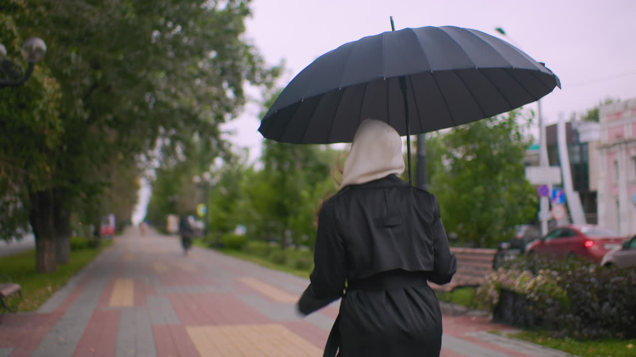 Back view of woman with white hood and black coat running under black umbrella along city sidewalk on cloudy rainy day, cars and trees lining street, autumn leaves scattered