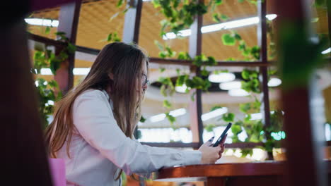 side view of woman in restaurant decorated with shopping bags on both sides operating her phone while her hair covers her face, capturing a cozy atmosphere in a modern dining space