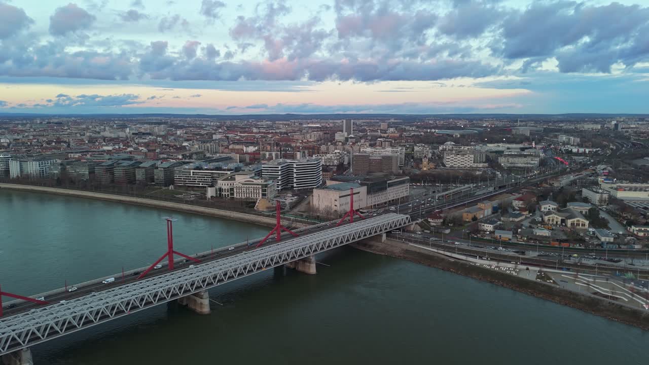 Vue aérienne orbitale de Budapest au coucher du soleil, soulignant le pont Rákóczi sur le Danube, entourée d'architecture urbaine, de trafic et de sites emblématiques comme le Müpa et le Théâtre National.
