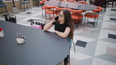 lady seated at table in restaurant making call, shopping bag beside her, surrounded by empty seats, creating a peaceful dining atmosphere