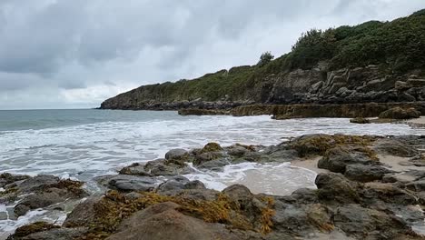 Slow-motion-ocean-waves-washing-over-rocky-coastal-shoreline-with-rugged-bay-cliff-across-the-skyline