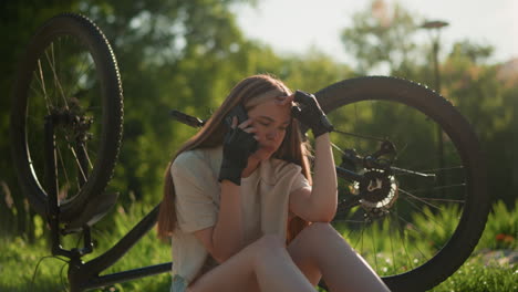 joven adulta sentada cerca de su bicicleta al revés, haciendo una llamada telefónica con una expresión preocupada y preguntosa, rodeada de árboles y plantas verdes vibrantes en un ambiente al aire libre sereno