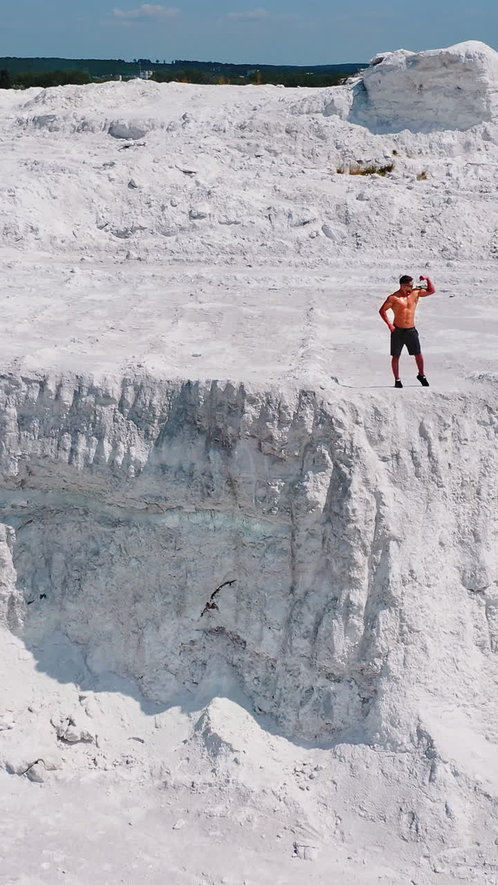 Muscular sportsman on a white hill. View from above on a strong guy who is showing his strength on the natural background. Bodybuilder on a rocky canyon. Aerial view. Vertical video