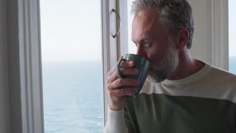 Caucasian-mature-man-looking-through-the-window-and-drinking-coffee