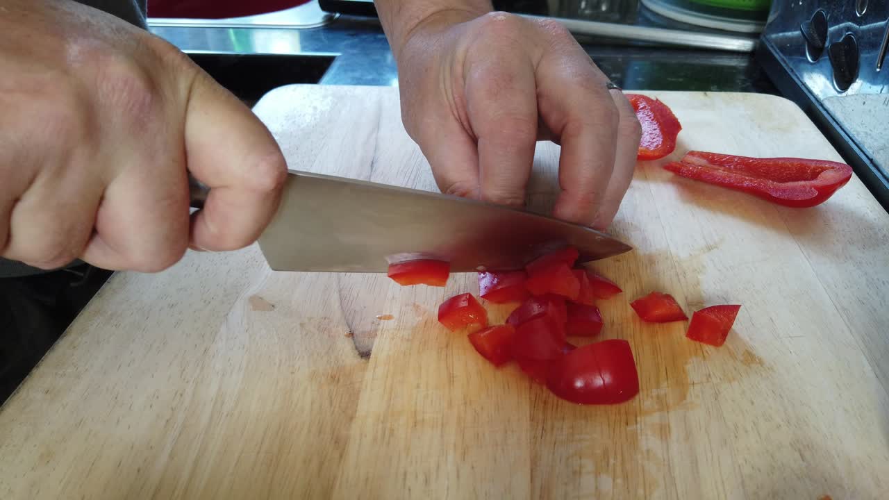 A man dices red peppers on a wooden chopping board at home.