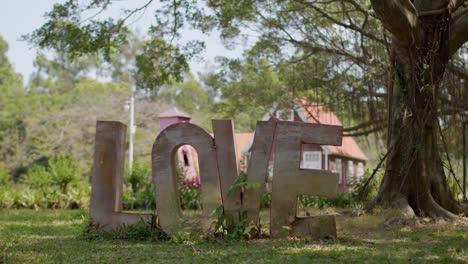 Letras-De-Metal-Que-Forman-La-Palabra-Amor-De-Pie-A-La-Sombra-De-Un-árbol-Cerca-De-Una-Casa-De-Estilo-Alemán-En-Segundo-Plano-En-Un-Día-Soleado