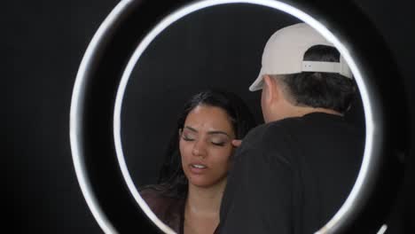 a makeup artist in a baseball cap does makeup on a blackhaired model under a lamp a makeup artist in a baseball cap does makeup on a blackhaired model under a lamp high quality footage