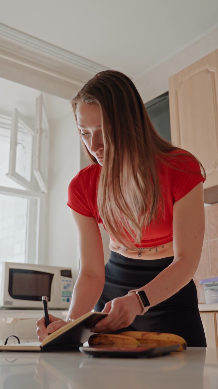 Una donna annota ricette per un'alimentazione sana, prepara spuntini monitorando tempo e nutrizione. Nella sua cucina, scrive piani alimentari e controlla le scorte per mantenere una buona salute