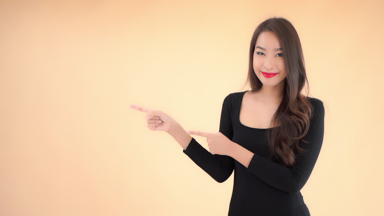 Attractive asian female with long hair in black dress pointing with fingers on left side, static full frame slow motion