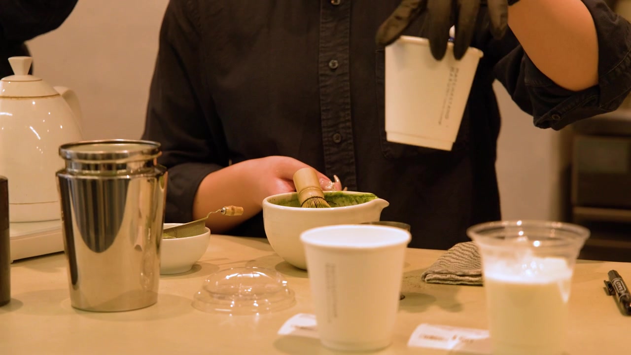 Close-up of a barista in black gloves preparing matcha green tea lattes in paper cups.