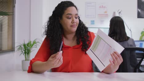 Happy-biracial-businesswoman-sitting-at-desk,-holding-documents,-making-video-call-in-modern-office