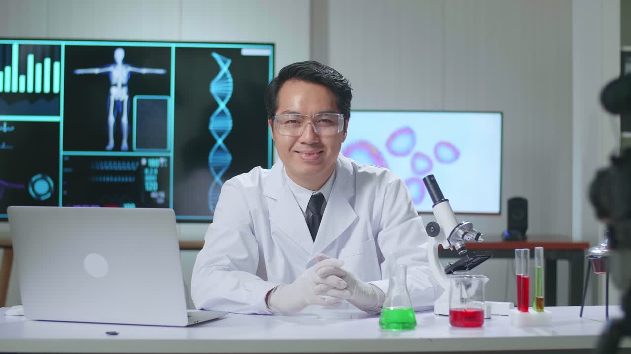 Asian Scientist Researching In The Laboratory With A Microscope, He Warmly Smiles Into The Camera