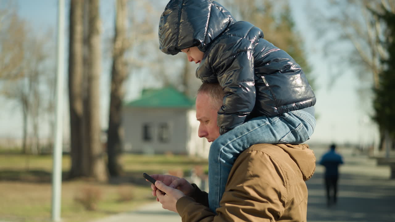 близкий план отца, несущего своего ребенка на плечах, когда он управляет своим телефоном, повернутый вправо, с некоторыми, идущими вниз, с деревьями и зданием позади