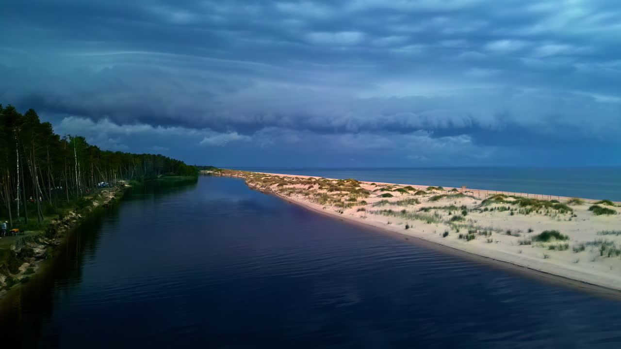 Pool of calm water reflects tall trees on edge of beach with sandy dunes and shallow grass, aerial ascend