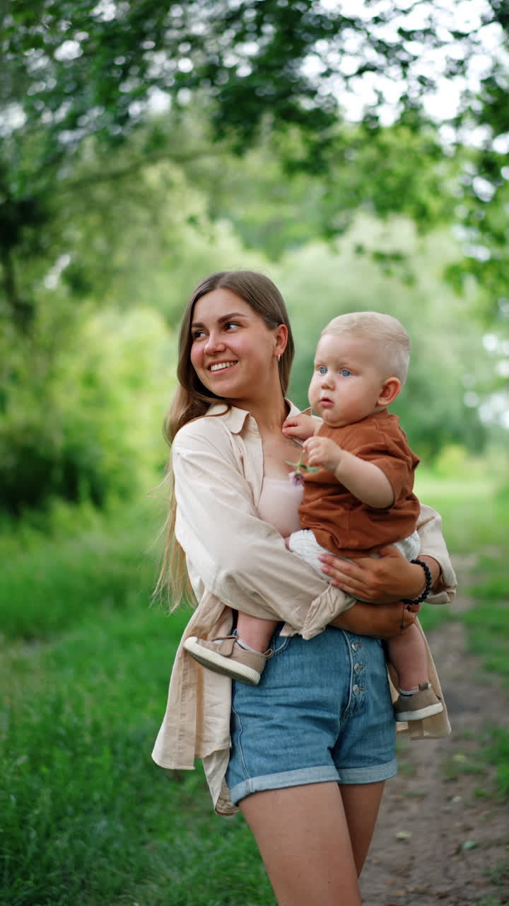 Long-haired Caucasian woman holding her baby in hands. Kid with flower in his hands looks around at nature with interest. Vertical video.