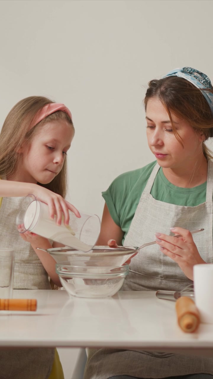 Woman and young girl collaborate in baking activity pouring ingredients into glass bowls on white table. scene features kitchen tools like rolling pins and whisks as they engage together